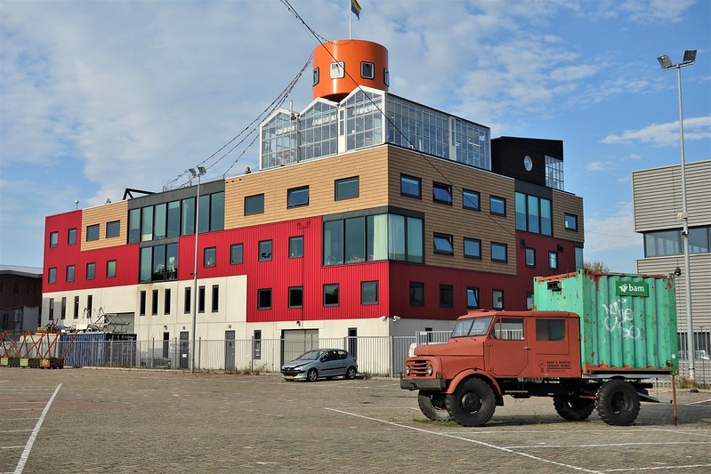 Een kleurrijk, modern gebouw met rood, beige en zwarte gevels en een oranje torentje bovenop. Het gebouw heeft grote ramen en lijkt een creatieve of industriële functie te hebben. Op de voorgrond staat een oude, roestige vrachtwagen met een groene container erop, die een industrieel en verweerd uiterlijk heeft. Het terrein rondom is grotendeels leeg, op een enkele geparkeerde auto na. Het contrast tussen het moderne gebouw en de oude vrachtwagen creëert een interessant beeld van vernieuwing versus verval.