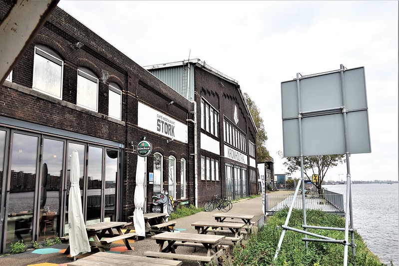 Een oud, industrieel gebouw aan het water, met de naam "Café-Restaurant Stork" op de gevel. Voor het gebouw staan houten picknicktafels en enkele opgevouwen parasols, wat duidt op een terras voor bezoekers. Het gebouw heeft grote ramen en een donkere bakstenen gevel. Op de achtergrond loopt een smal pad langs de waterkant, met een hek dat het scheidt van het water. Fietsen zijn geparkeerd tegen de gevel, en de sfeer is rustig en uitnodigend, ideaal voor een ontspannen bezoek aan het water.