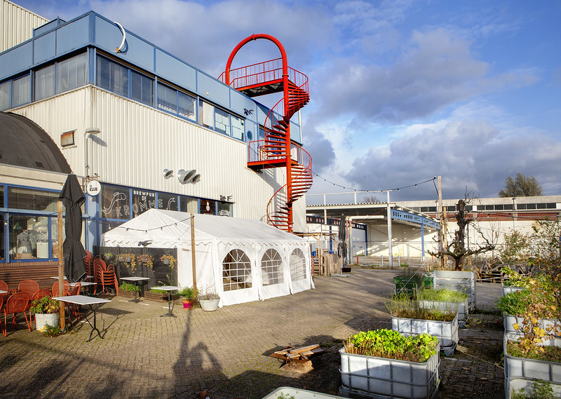 Tent met ramen, een terras met tafels en stoelen, en een opvallende rode wenteltrap die naar het dak leidt. De omgeving heeft een industriële uitstraling met grote plantenbakken en verschillende decoraties. De lucht is deels bewolkt, wat een frisse sfeer geeft.