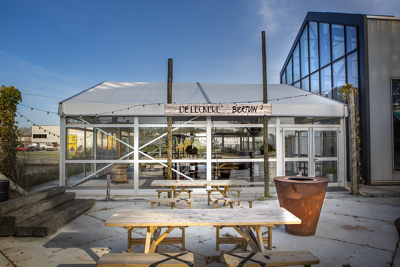 Een zonnig terras met houten picknicktafels bij de 'De Leckere Biertuin'. Het terras is gelegen naast een glazen serre-achtig gebouw en is versierd met lichtsnoeren. Op de achtergrond staat een bord met de naam van de biertuin.