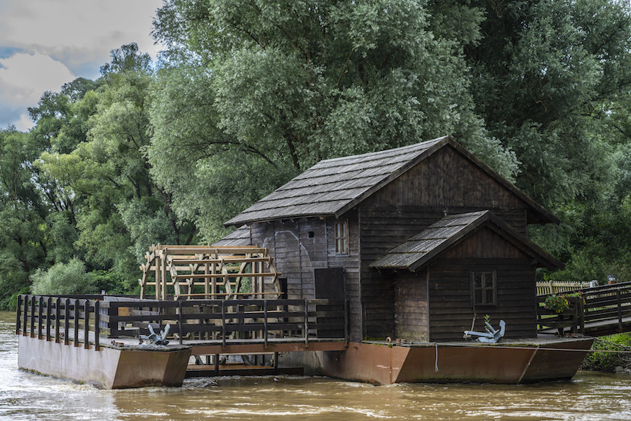 Foto van een donkerhouten watermolen aan een groenige rivier met bomen op de achtergrond.