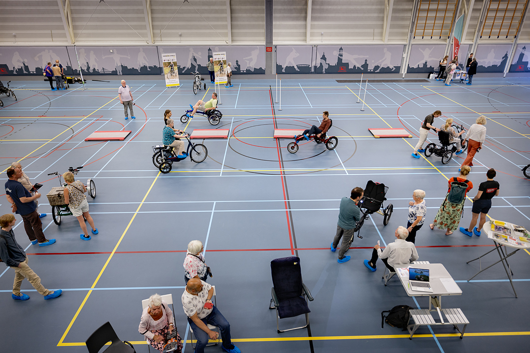 In een gymzaal (Europahal) proberen senioren driewielfietsen uit. Er liggen matten op de grond waar ze overheen kunnen fietsen. En er staat begeleiding op de foto die hen helpen.