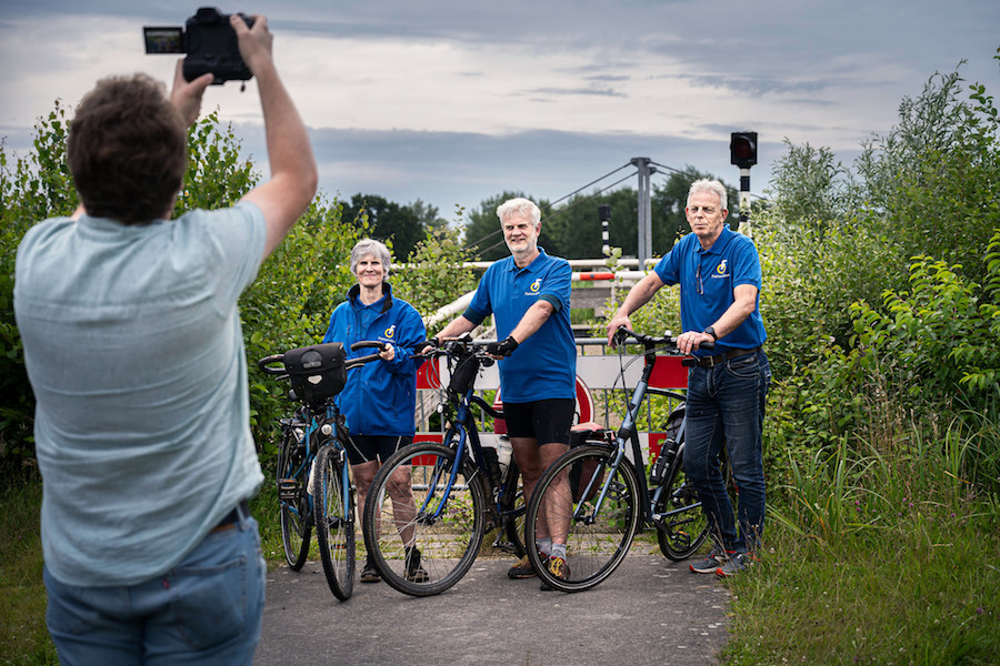 Een man fotografeert drie fietsers in blauwe shirts met Fietsersbondlogo voor een afgesloten brug in Giethoorn.