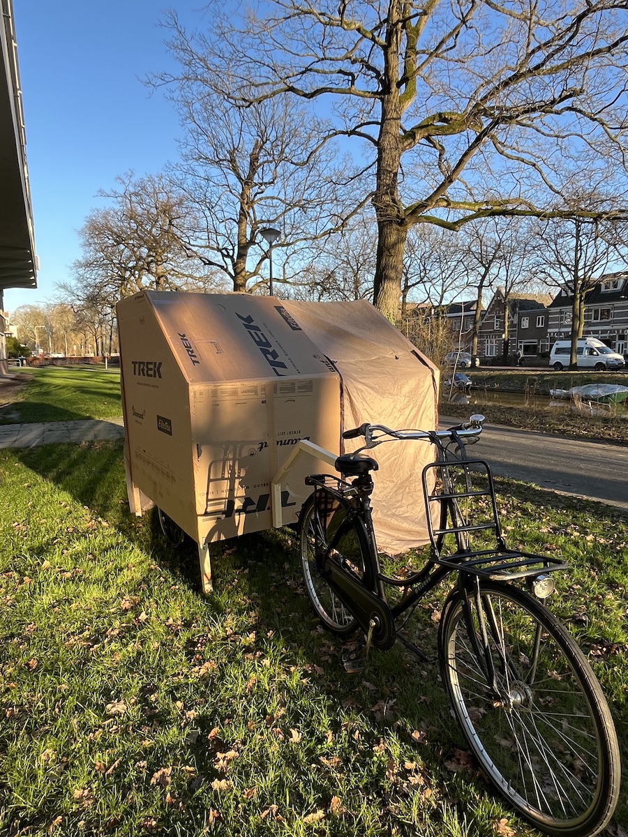 Fietscaravan staat achter de fiets. Pootjes zijn al omlaag. Hij is klaar om geinstalleerd te worden als minihuisje.