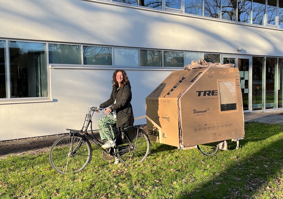 Vrouw met lange donkere haren heeft achter haar fiets een kartonnen fiestcaravan