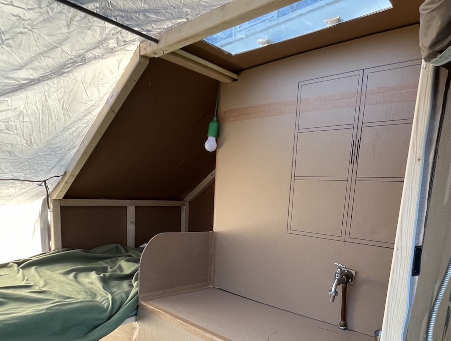 Het interieur van de van de mobiele studentenwoning met op karton kastjes getekend en daarvoor een echte kraan. Links zie je nog een matras liggen met groene matrasovertrek.