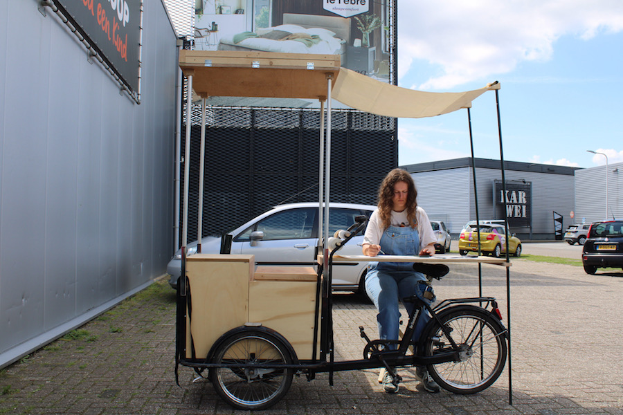 Meisje met lange haren zit te schetsen aan een tafelblad op een bakfiets met luifel boven zich. Omgeving: industrieterrein.