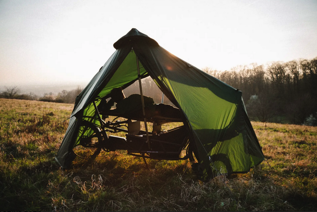 Man ligt op een stretcher onder een tentdoek die in vorm wordt gehouden door een fiets. Omgeving: ochtendlicht op weide.