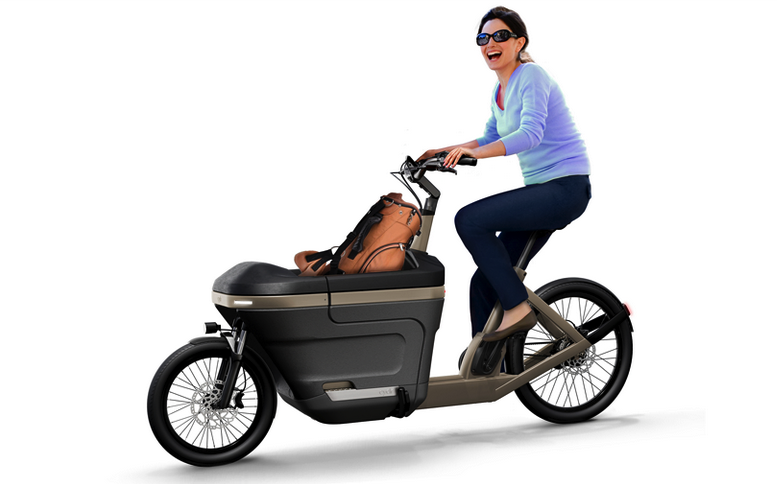 Vrouw met zonnebril op zit schaterend op een elektrische bakfiets met daarin een lichtbruine tas