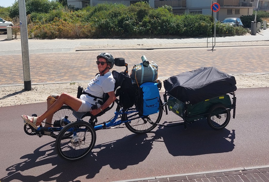 Annette Verberne Rijdt met haar elektrische driewielfiets en haar fietskar met opgevouwen tent door een zonnige straat.