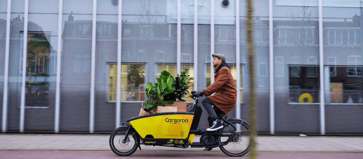 Man met pet en bruine jas rijdt op een gele bakfiets met grote plant in de bak. Op de bak staat in zwarte letters 'Cargaroo'.