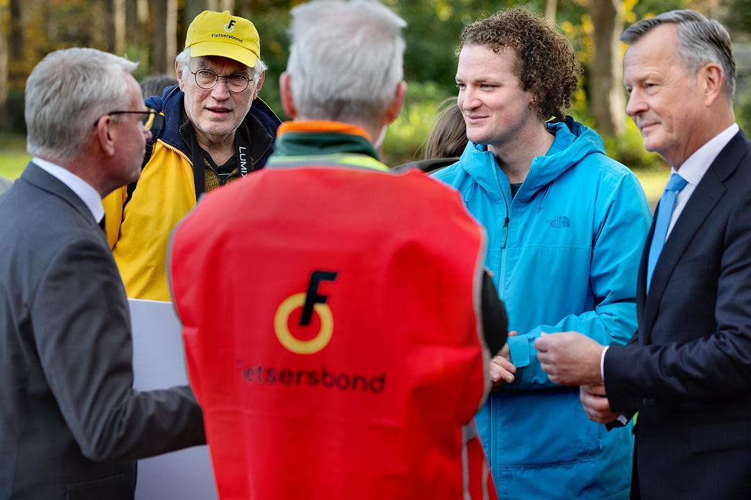 Vrijwilliger van de Fietsersbond heeft een rood hesje aan en staat naast een andere vrijwilliger, Ross Goorden van de Fietsersbond en de gedeputeerde en CvdK.
