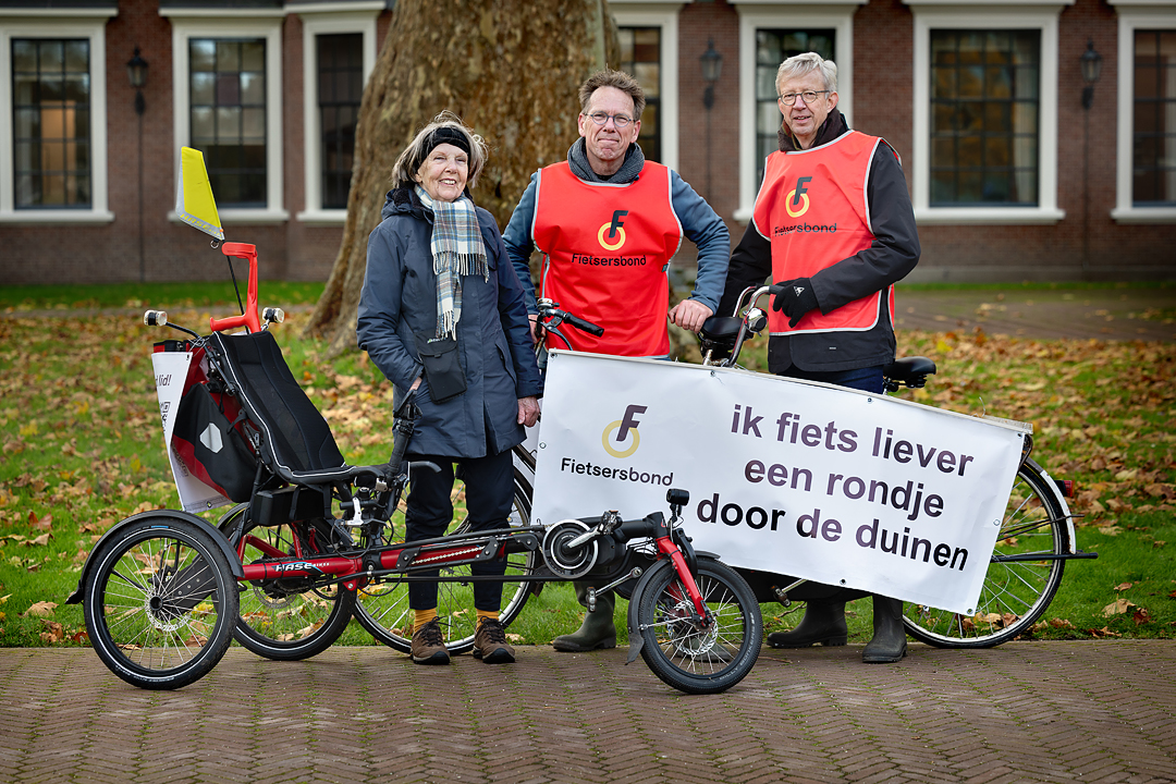 Drie personen staan naast een handbike en een tandem met een spandoek met de tekst: Ik fiets liever een rondje door de duinen. Ze voeren actie voor het behoud van de duinpaden en twee hebben een Fietsersbond hesje aan.