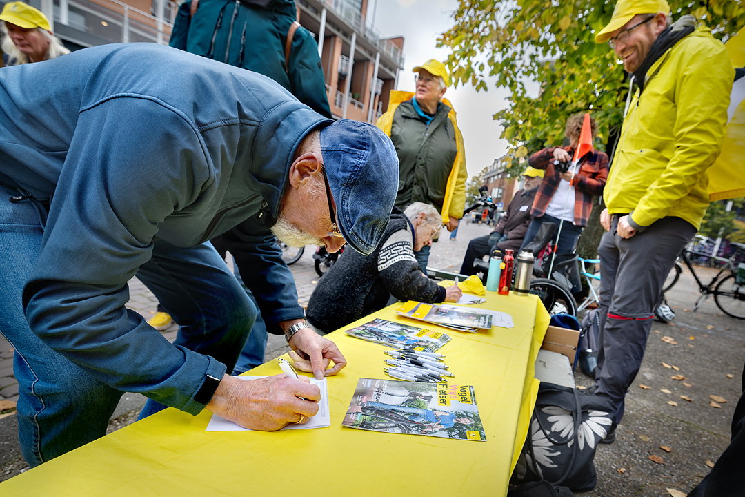 Een man zet een handtekening onder de petitie fietsparkeren van de Fietsersbond. Om hen heen staan vrijwilligers van de Fietsersbond met een gele pet op.