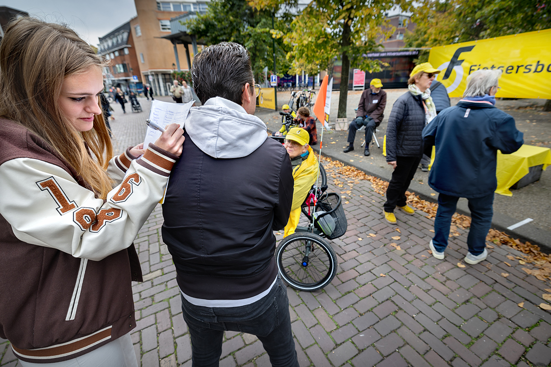 Een vrouw zet een handtekening onder de petitie fietsparkeren van de Fietsersbond. Ze houdt de petitie op de rug van een man om hem te kunnen ondertekenen. Om hen heen staan vrijwilligers van de Fietsersbond met een gele pet op.