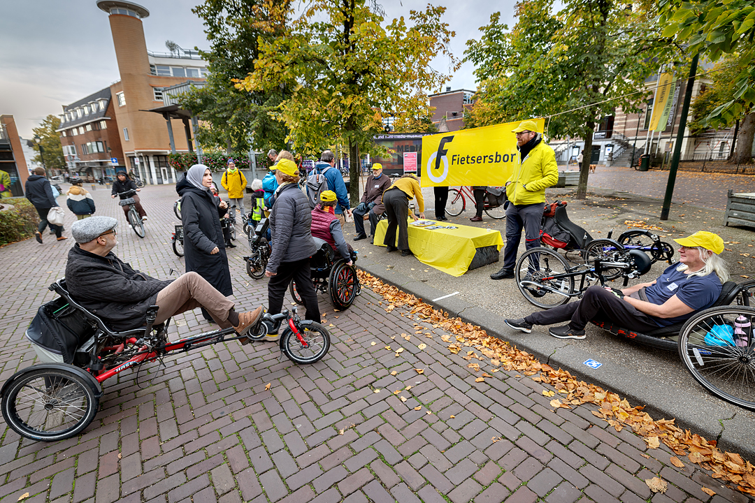 Naast een geel spandoek staat een lage tafel met een geel laken vol petities en pennen. Een vrijwilliger van de Fietsersbond praat met omstanders. Om hen heen zitten vrijwilligers van de Fietsersbond op een handbike of driewielfiets.