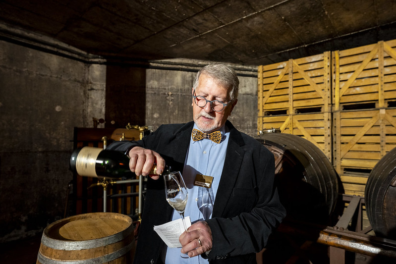 man met vlinderdas schenkt een glas wijn in met op de achtergrond kisten en een wijnton.