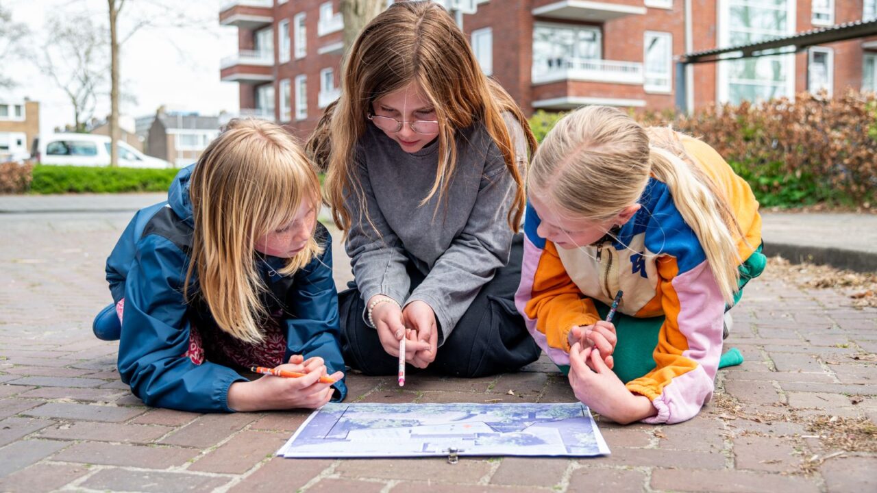 Drie meisjes zitten gehurkt op straat met een pen in de hand en kijken naar een kaart van hun schoolomgeving.