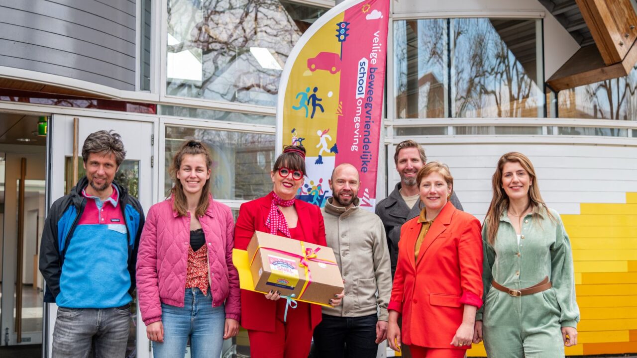 Zeven volwassenen staan voor de ingang van een basisschool met een kartonnen box in hun handen: de eerste versie van de Challengesbox.