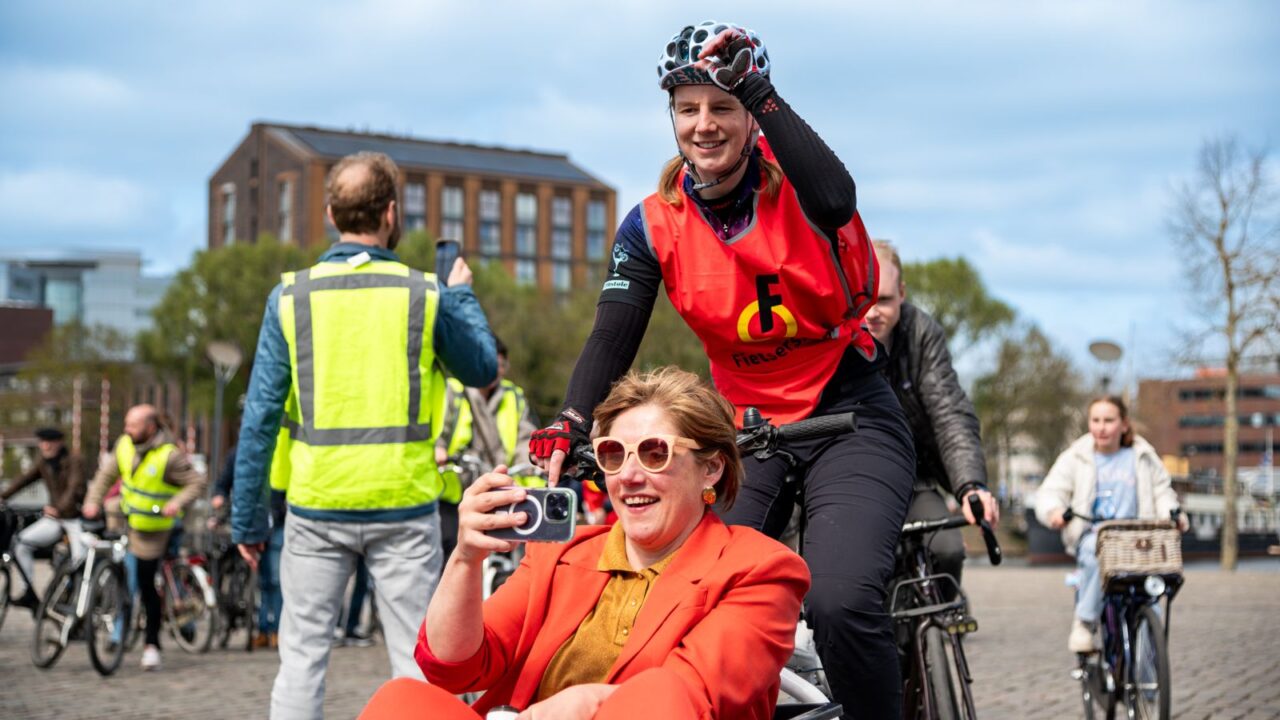 Vrouw zit voorop in bakfiets en maakt foto's met haar telefoon. Ze wordt voortgetrapt door een vrouw met een Fietsersbond hesje aan. Op de achtergrond meer volwassenen en kinderen op de fiets.