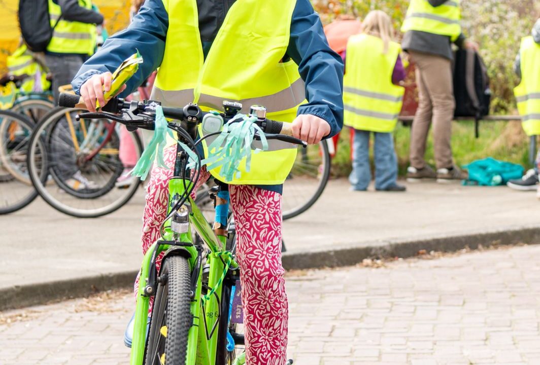 Meisje met geel hesje aan op haar versierde fiets. Op de achtergrond zijn meer kinderen en volwassenen met een geel hesje aan te zien.