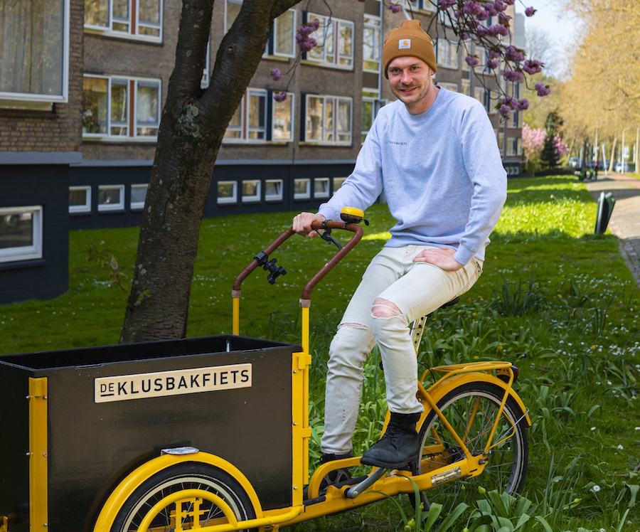 Jonge man met okerkleurige beanieop gele bakfiets met daarop: De Klusbakfiets.