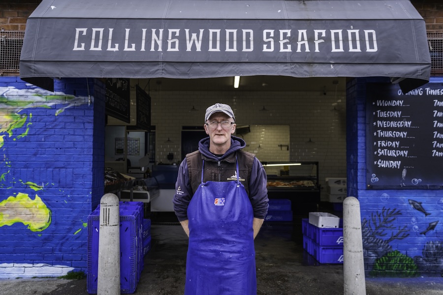 Man met blauw plastic schort voor staat voor luifel met daarop 'Collingwood seafood'.