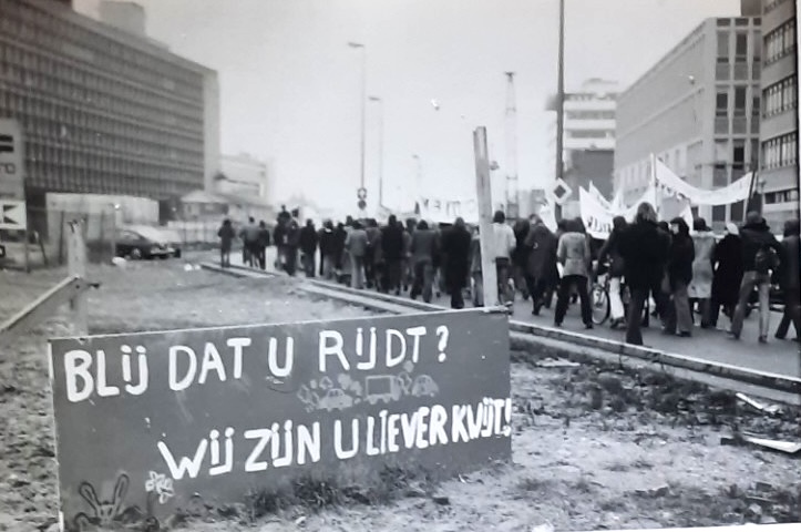 Bord met de tekst: 'blij dat u rijdt?' op een open stuk asfalt waarnaast een stoer demonstranten over de weg loopt.