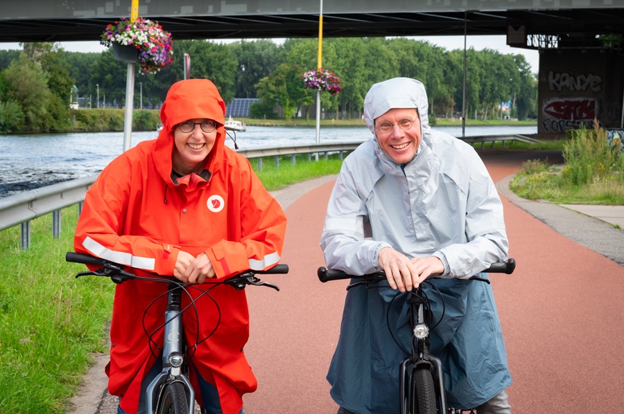 Vrouw met knaloranje pocho en man in lichtblauwe capuchon met hun fietsen langs een kanaal.