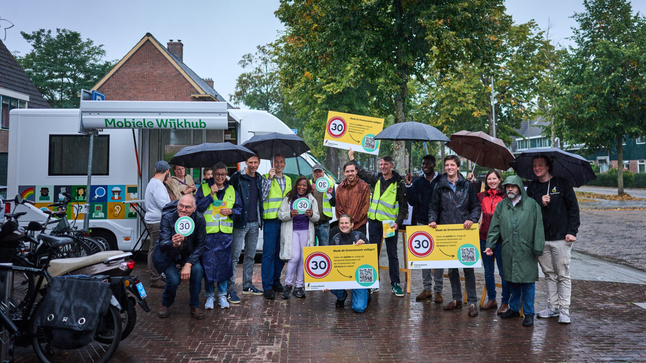 Leden van de Fietsersbond en buurtbewoners staan met petitieborden en 30 km/u borden op straat. Vanwege de regen met paraplu's in de hand.