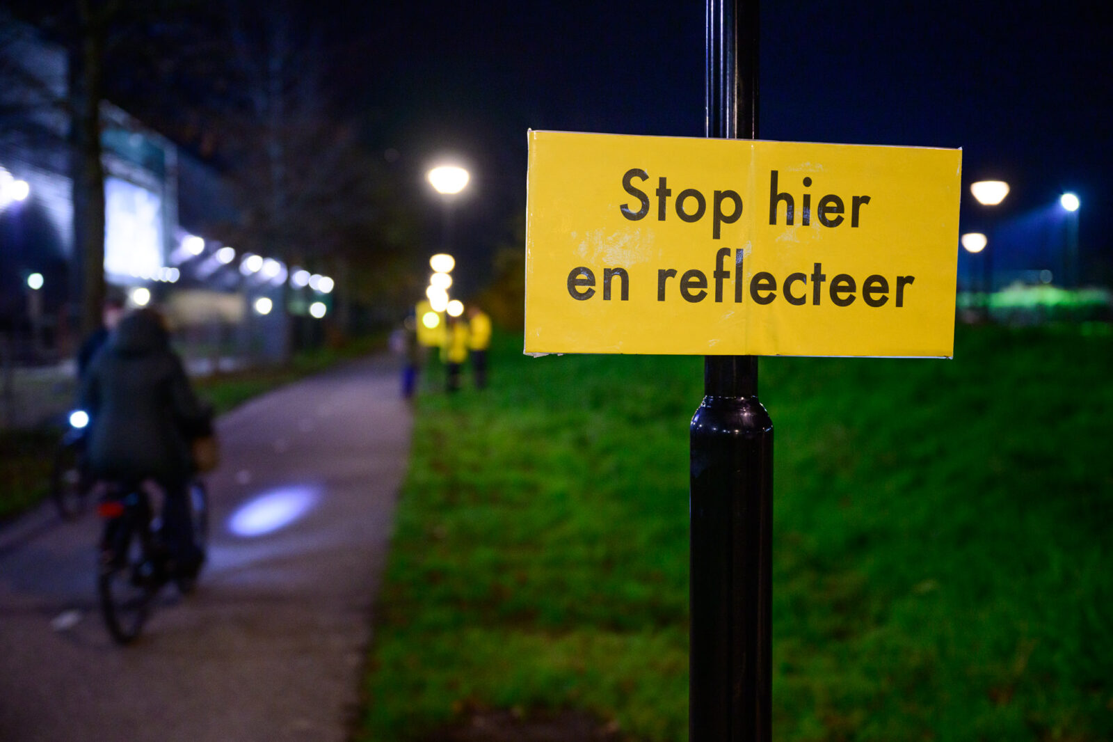 Bordje 'Stop hier en reflecteer' op een paal langs een donker fietspad.