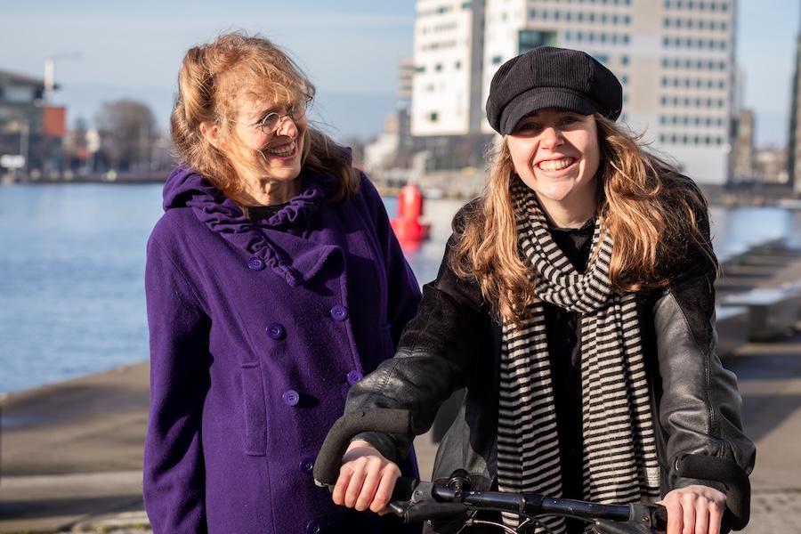 Lachende vrouw in paarse jas kijkt naar vrouw met pet en fiets met op de achtergrond het IJ in Amsterdam.