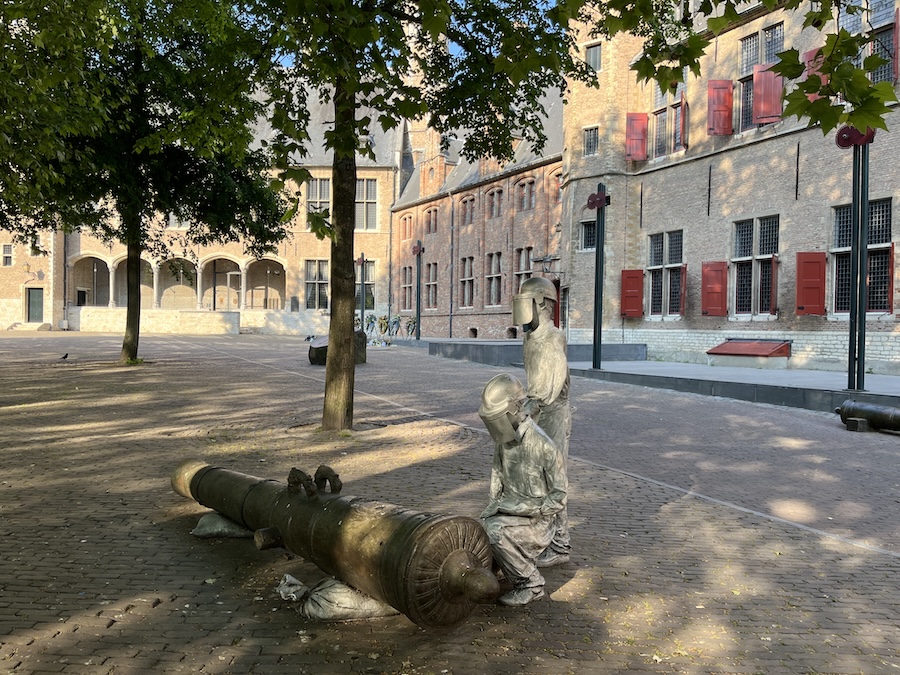 Twee zilverkleurige figuren staan bij de gele vrom van een kanon op een groot plein met oude hoge gebouwen met rode luiken.