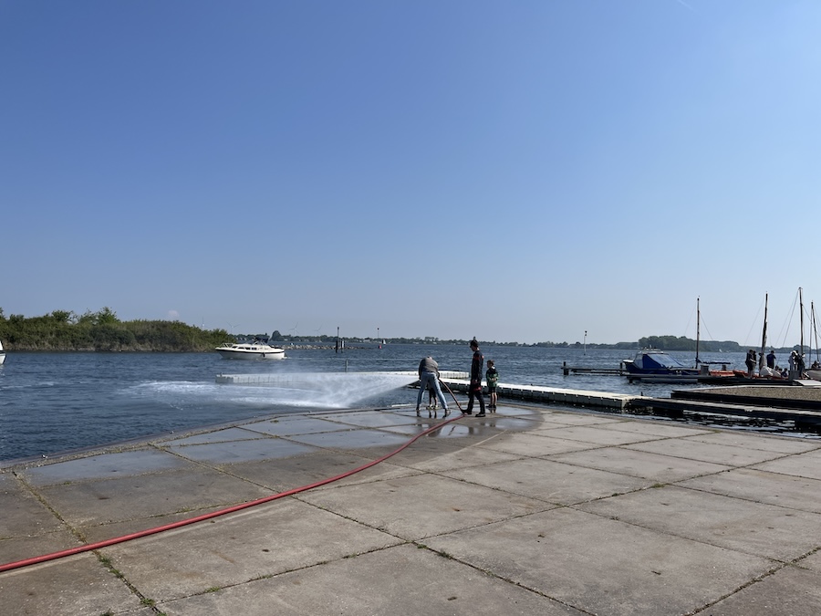 Op een werf spuit een brandslang op het water met rechts kleine bootjes.