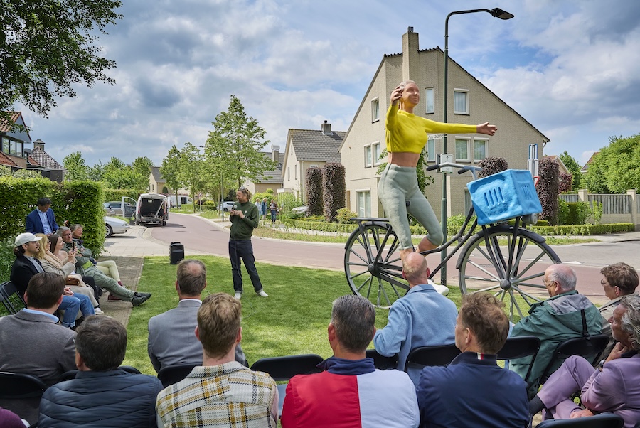 Groep mensen staat rondom een groot beeld van een meisje met paardenstaart die op de fiets zit en haar armen van hte stuur houdt.