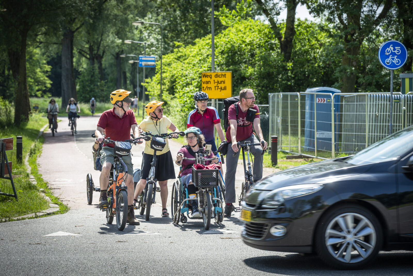 Een groep fietsers, van vouwfiets tot driewielfiets, wacht voor een passerende auto