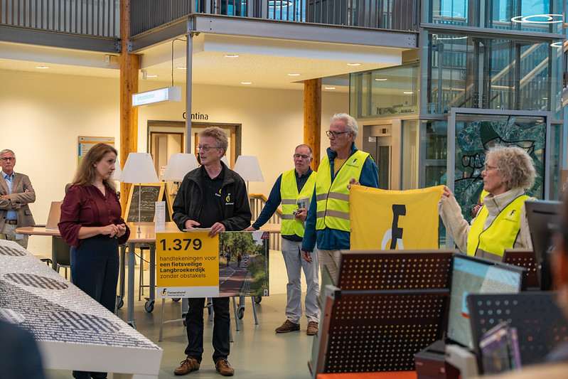 Groep mensen staan in een bibliotheek met vlaggen en borden om petitie te overhandigen aan