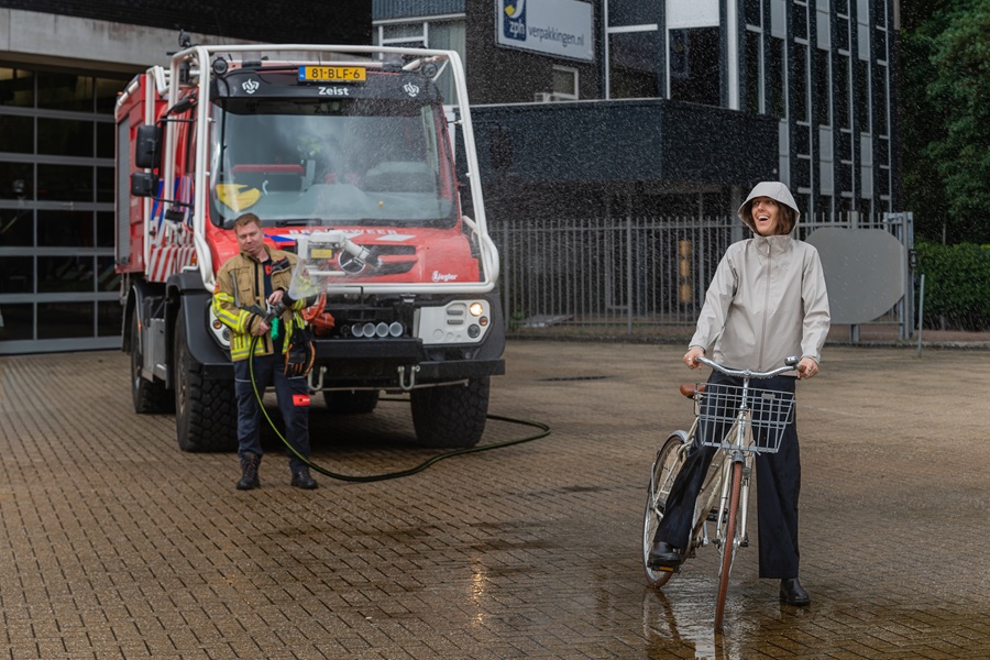 Brandweerman zet spuit op lachende fietser in regenjas met capuchon op.