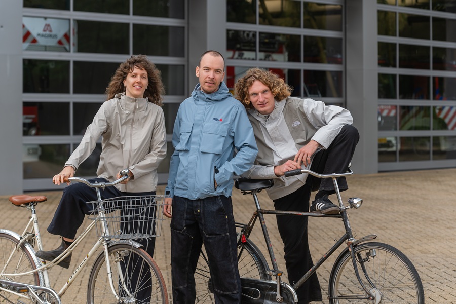 Drie fietsers in regenjassen staan naast elkaar voor ramen waarachter je een brandweerauto ziet.